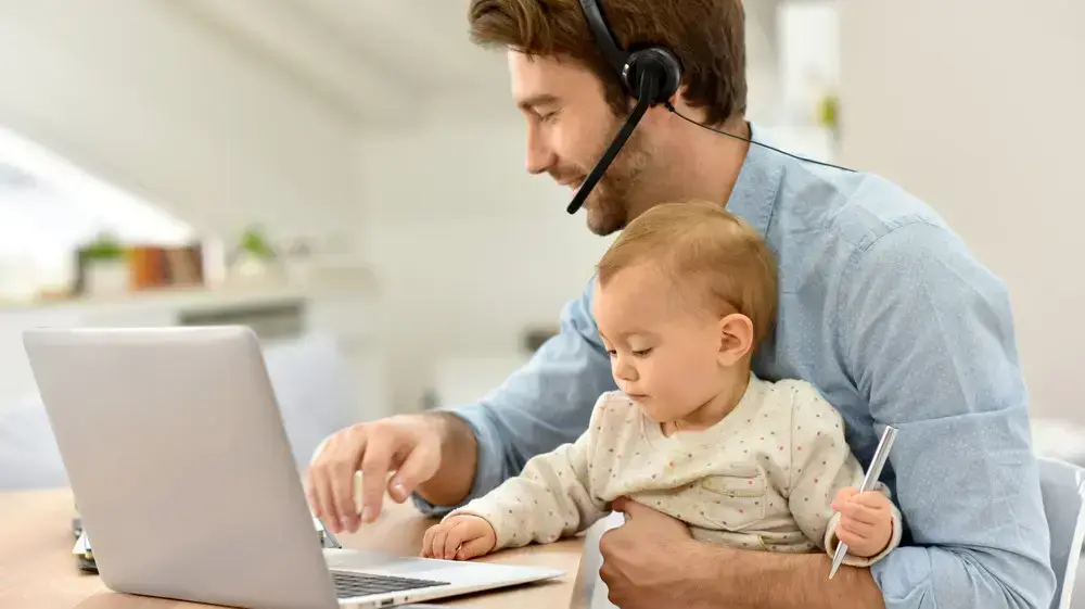 גבר יושב מול לפטופ עם אוזניה ותינוקת על הברכיים גבר יושב מול לפטופ עם אוזניה ותינוקת על הברכיים
