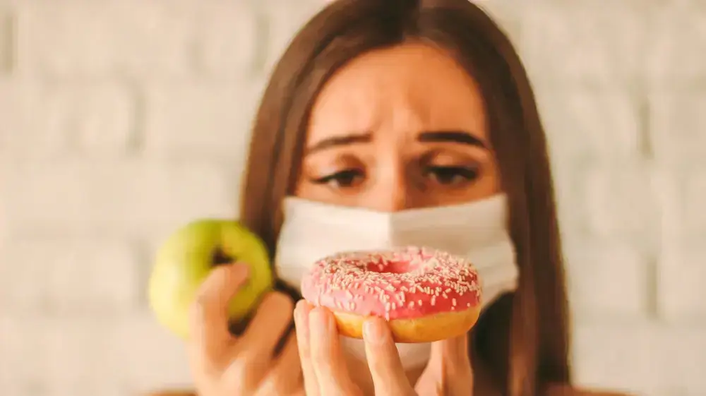 אישה במסיכה מתלבטת בין דונאט לתפוח אישה במסיכה מתלבטת בין דונאט לתפוח