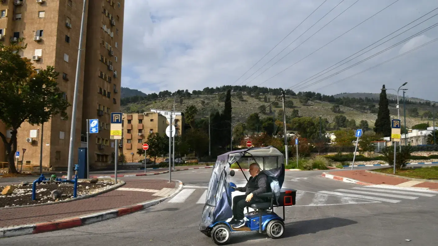 גבר נוסע על קלנועית וברקע דגל ישראל, קריית שמונה, 2 בינואר 2024 גבר נוסע על קלנועית וברקע דגל ישראל, קריית שמונה, 2 בינואר 2024