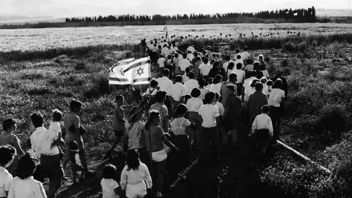קבוצת בני נוער עם דגלי ישראל וחרמשים, בדרכה לשדה לחיתוך הראשון של שעורה בערב של היום הראשון של פסח. סביבות 1960.