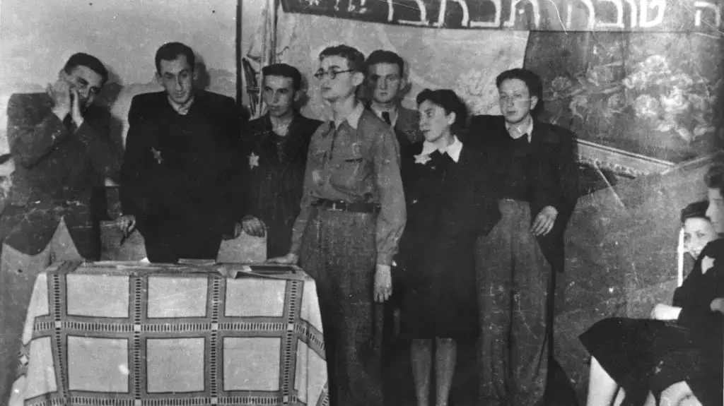 גלויות יד ושם לכתבה של אור רביד, צילום מסיבת ראש השנה של ארגון חזית דור בני המדבר, לודז' פולין, 1942. הקבוצה לבושה בגדי חג ועוטים טלאי צהוב. צילם: מנדל גרוסמן. גלויות יד ושם לכתבה של אור רביד, צילום מסיבת ראש השנה של ארגון חזית דור בני המדבר, לודז' פולין, 1942. הקבוצה לבושה בגדי חג ועוטים טלאי צהוב. צילם: מנדל גרוסמן.